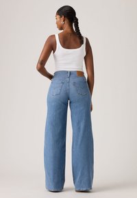 Τα ανοιχτό μπλε τζιν με φαρδιά μπατζάκια διαθέτουν ψηλή μέση, πίσω τσέπες και καφέ δερμάτινο λογότυπο. Το μοντέλο φοράει λευκή αμάνικη μπλούζα.