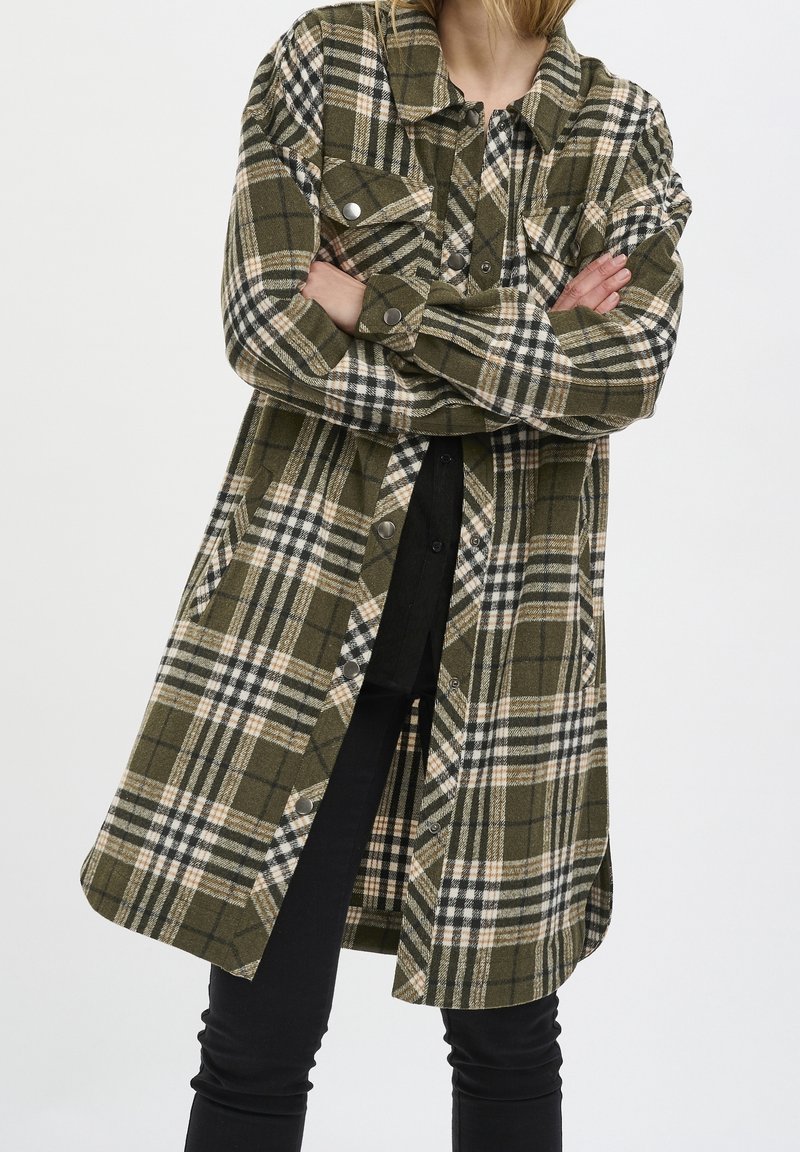 Veste chemise à carreaux en vert olive, crème et noir, avec fermeture à boutons, coupe oversize, poches et légère évasement à l'ourlet.