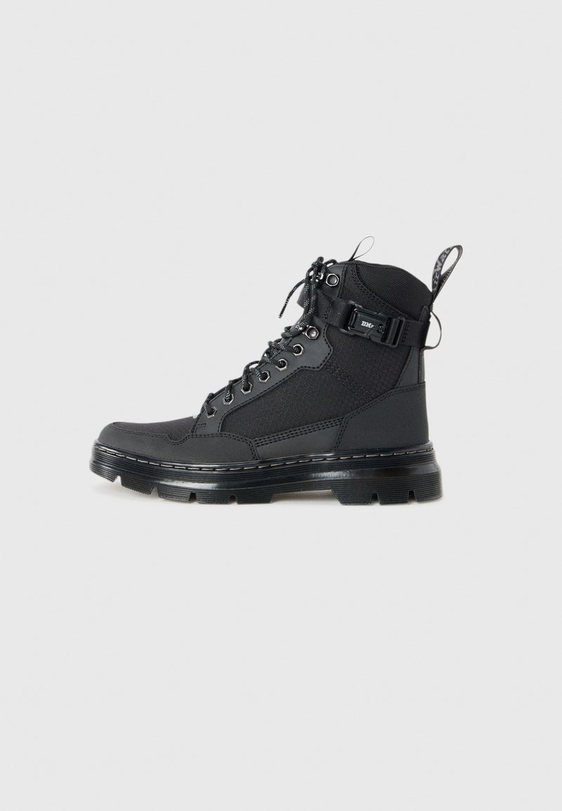 Botte de combat noire à lacets avec semelle épaisse, bride de cheville avec boucle, dessus en tissu et languette sur fond gris clair.