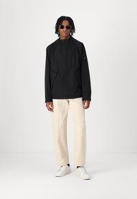 Schwarze wasserdichte Jacke mit Kapuze, zwei Taschen vorne und einem Logo am linken Ärmel, kombiniert mit cremefarbigen Hosen und weißen Turnschuhen.
