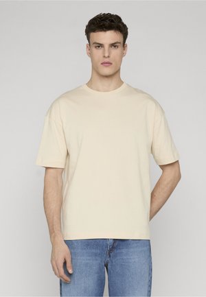 Giovane uomo con capelli corti e ricci che indossa una t-shirt beige tinta unita a maniche corte con collo a giro e jeans blu, in piedi davanti a uno sfondo grigio.