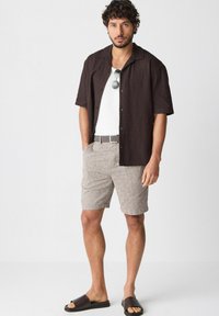 Brun kortærmet skjorte med knaplukning og et tekstureret mønster, hvid top, lysebeige shorts og brune sandaler. Solbriller på skjorten.