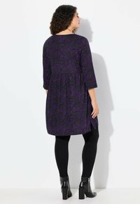 Robe tunique à motifs floraux violets en tissu doux, dotée de manches trois-quarts, d'une taille froncée et de fentes latérales. Portée avec des leggings noirs.