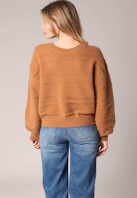 Femme aux cheveux blonds portant un pull brun loose et texturé, avec un jean bleu clair, de dos contre un fond beige uni.