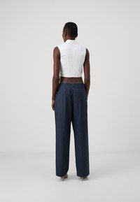 Femme portant un débardeur blanc sans manches et un pantalon large rayé bleu marine, se tenant de dos sur un fond uni.