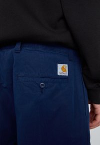 Pantalones azul marino con un bolsillo trasero que cuenta con un cierre de botón, acompañados de una etiqueta blanca cuadrada con un logotipo naranja. Material de algodón.