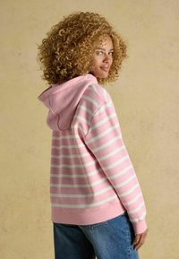 Žena s kučeravými vlasmi, oblečená v ružovo-bielom pruhovanom mikinovom saku a modrých džínsoch, stojí bokom proti béžovému pozadiu.