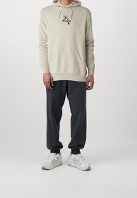 Persona che indossa un hoodie beige con una libellula e il testo "New Era", pantaloni jogger grigio scuro e sneakers bianche, in piedi contro uno sfondo semplice.