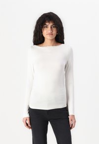 Long-sleeve, off-white top made of stretchy, smooth fabric with a boat neckline and gathered detail at the sides. Paired with black pants.
