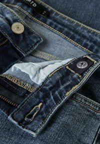 Jean en denim bleu foncé avec des bords effilochés, mettant en avant un tissu intérieur bleu clair et des fermetures à bouton en argent. Accents cousus le long des coutures.