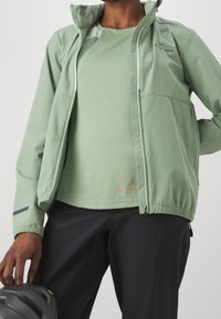 Vaaleanvihreä vetoketjullinen takki, jossa on joustavat hihansuut ja kaulus. Pinnaltaan sileä ja siinä on heijastava yksityiskohta. Päällä samanvärinen yläosa.