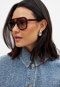 Brown oversized sunglasses with a geometric frame, worn with a textured blue denim jacket. Silver teardrop earrings are also visible.