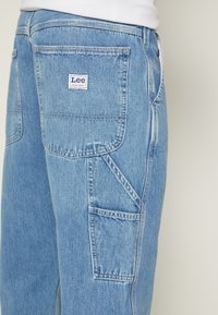 Blå denim arbetsbyxor med rak skärning, prydda med en sydd etikett på bakfickan och flera praktiska fickor.