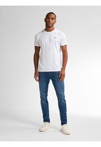 Camiseta blanca de cuello redondo hecha de algodón, combinada con jeans azules y zapatillas blancas. La camiseta tiene un pequeño logo en el pecho.