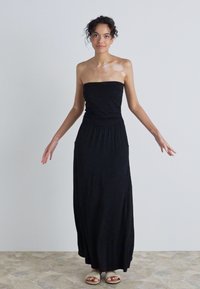 Svart strapless maxikjole med en tettsittende bodice og samlet midje. Lett stoff; glatt tekstur. Modell iført sandaler, stående mot en nøytral bakgrunn.