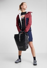 Zaino impermeabile nero con una superficie testurizzata, dotato di un logo grigio. Modello che indossa una giacca bordeaux e blu, una maglietta nera e pantaloni corti.