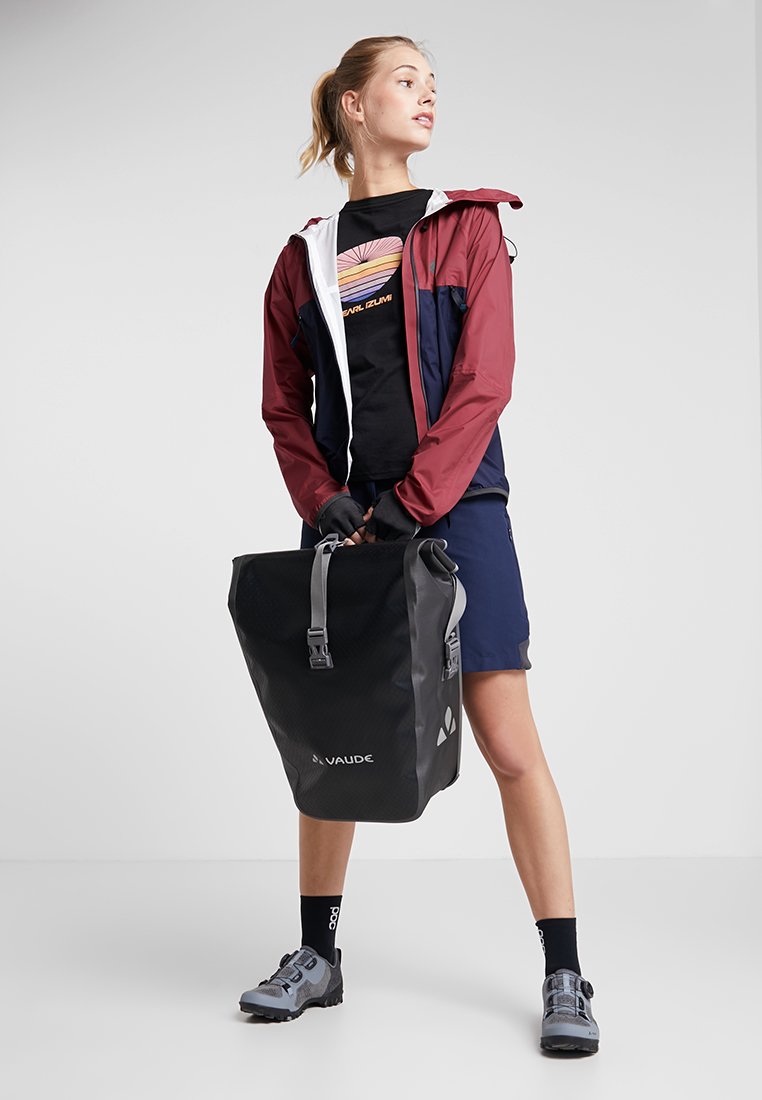 Zaino impermeabile nero con una superficie testurizzata, dotato di un logo grigio. Modello che indossa una giacca bordeaux e blu, una maglietta nera e pantaloni corti.