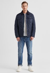 Marineblaue Steppjacke mit Wollkragen, ausgestattet mit Vorderfachern und Druckknöpfen, getragen über einem weißen Hemd und blauen Jeans mit aufgerollten Bündchen.