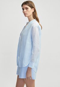 Hellblaue Leinen-Hemdbluse mit langen Ärmeln, runder Säume und zarter Spitzendekoration am Kragen. Kombiniert mit passenden Shorts.