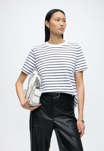 Femme aux longs cheveux noirs portant une chemise blanche à rayures et un pantalon noir, tenant un petit sac à main blanc sur un fond uni.