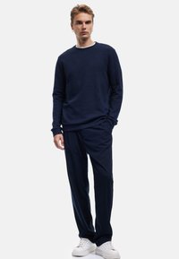 Maglione blu navy con texture e design a coste, abbinato a pantaloni blu navy e sneakers bianche, in piedi contro uno sfondo neutro.