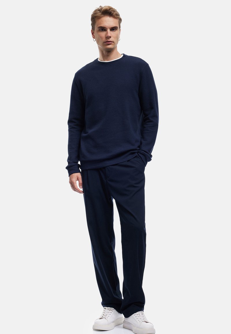 Maglione blu navy con texture e design a coste, abbinato a pantaloni blu navy e sneakers bianche, in piedi contro uno sfondo neutro.