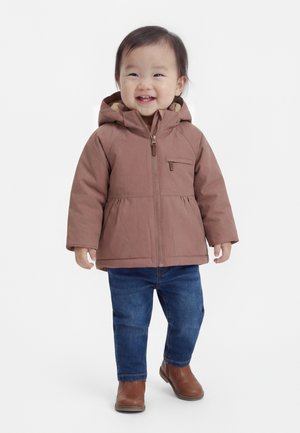 Bambin souriant, portant une veste à capuche marron, un jean bleu et des bottines marron, debout sur un fond blanc.