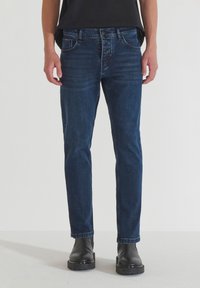 Jeans de mezclilla azul oscuro con corte ajustado, diseño de cinco bolsillos y detalles de costura sutiles, combinados con botas negras hasta el tobillo.