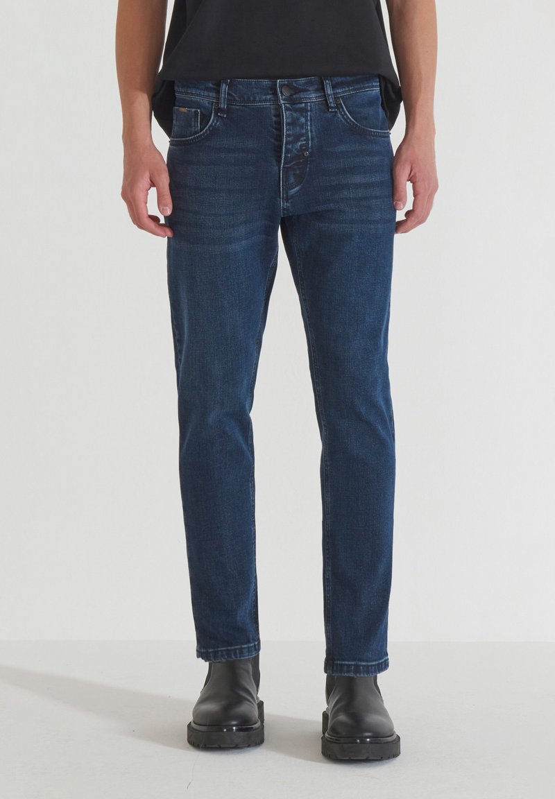 Jeans de mezclilla azul oscuro con corte ajustado, diseño de cinco bolsillos y detalles de costura sutiles, combinados con botas negras hasta el tobillo.