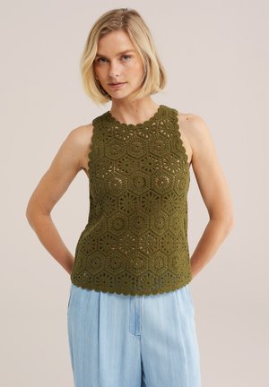 Nő olívazöld horgolt ujjatlan felsőben és világoskék nadrágban, háttal állva kezét a háta mögött tartva, egyszínű háttér előtt.