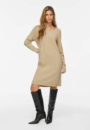 Femme debout portant une robe-pull beige à manches longues et des bottes noires montantes à talons sur un fond blanc uni.