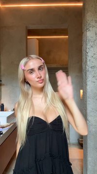 Svart strapless klänning med volangkjol, långt blont hår, två rosa hårklämmor. Inomhusmiljö med mjukt ljus och strukturerade väggar.