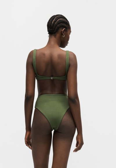 Femme aux cheveux tressés portant un maillot de bain deux pièces côtelé vert olive, vue de dos sur un fond clair uni.