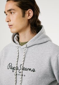 Sudadera gris de tejido suave con un logo frontal en impresión oscura, cordones con acentos estampados y un corte relajado.
