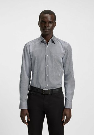Chemise à manches longues, à boutons, avec de fines rayures verticales bleues et blanches, col évasé, coupe slim et poignets réglables, portée avec un pantalon noir et une ceinture.