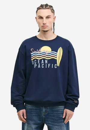 Mężczyzna w granatowej bluzie z grafiką deski surfingowej, słońcem i napisem „Surfing Ocean Pacific”, w połączeniu z niebieskimi dżinsami, stojący na białym tle.