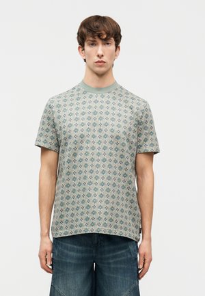 Νέος άνδρας που φοράει κοντομάνικο πουκάμισο με πράσινα και μπλε λουλουδάτα μοτίβα και σκουρόχρωμα μπλε τζιν σορτς, στέκεται μπροστά σε ένα απλό φόντο.