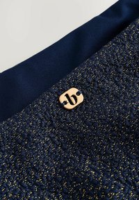 Marine stof met een gestructureerd patroon dat verweven is met gouden draden; afgewerkt met een ronde bronzen logo-accent.