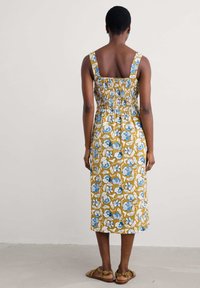 Bloemige zomerjurk in mosterd met blauw en witte patronen, voorzien van brede bandjes, een gerimpelde achterkant en een midi-lengte, gecombineerd met bruine sandalen.