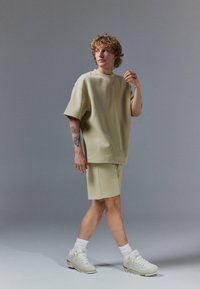 Jeune personne aux cheveux bouclés portant un T-shirt beige oversize, un short assorti, des chaussettes blanches et des baskets blanches, avec un bras tatoué visible.