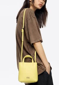 Żółta skórzana torebka z dwoma uchwytami i odpinanym paskiem na ramię, o ziarnistej fakturze i tłoczonym logo marki z przodu.