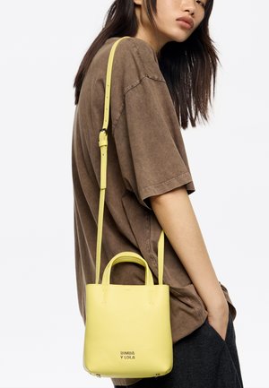 Bolso de cuero amarillo con dos asas y una correa de hombro desmontable, con textura granulada y logo de la marca en relieve en la parte frontal.