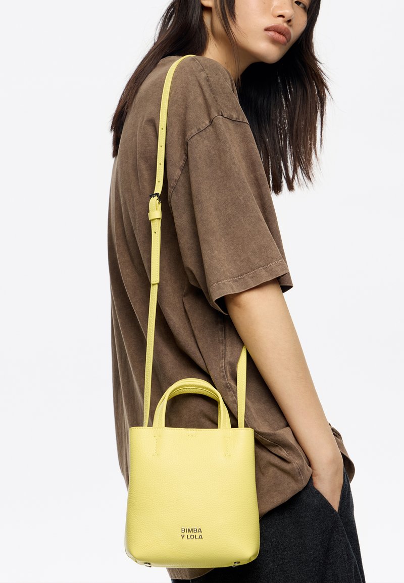 Żółta skórzana torebka z dwoma uchwytami i odpinanym paskiem na ramię, o ziarnistej fakturze i tłoczonym logo marki z przodu.