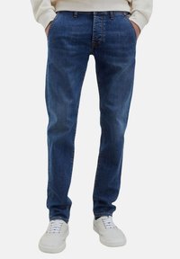 Jeans in denim medio lavato da uomo con taglio dritto, caratterizzati da cinque tasche e dettagli di cucitura sottili, abbinati a sneaker bianche.