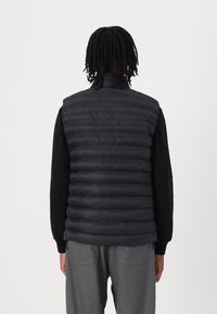 Black puffer vest with horizontal stitching, high collar, worn over a black long-sleeve shirt, paired with gray pants.