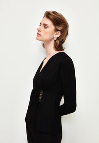 Top negro de manga larga con cuello en V, diseño estructurado y detalles en dorado en la cintura, con una silueta ajustada y textura suave.