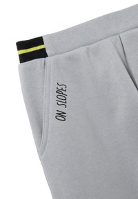 Pantalones de chándal gris claro con una cinturilla elástica a rayas en negro y amarillo. Presenta el texto "ON SLOPES" impreso en negro en el bolsillo lateral.