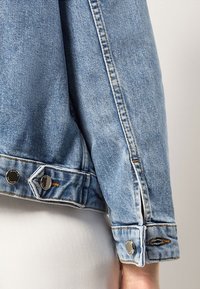 Hihaton vaaleansininen farkkutakki, jossa on hopeiset nepparit ja oranssia ommelta, henkilön käsivarren päällä yksinkertaista taustaa vasten.