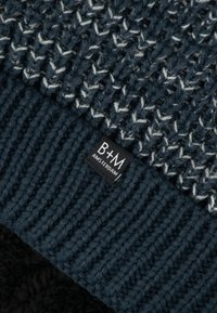 Dunkelblau gestrickte Mütze mit strukturiertem Muster und weißen Akzenten. Mit einem schwarzen B+M Amsterdam-Label am umgeschlagenen Rand.