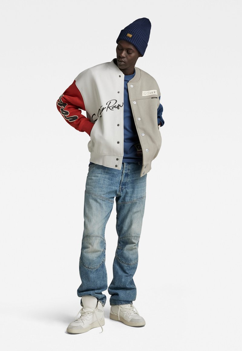 Varsityjack met een beige lichaam, rode en blauwe mouwen, met logo en tekstdetails. Gepaarde met lichtblauwe jeans en witte sneakers.